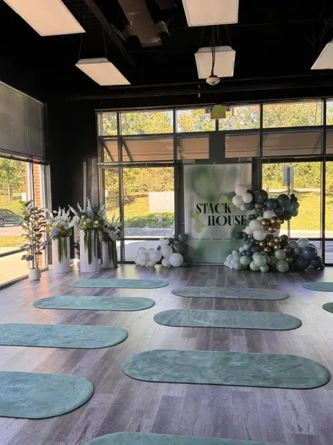Bright event space with white pedestal arrangements at left, a navy, sage and white balloon garland, and teal floor mats