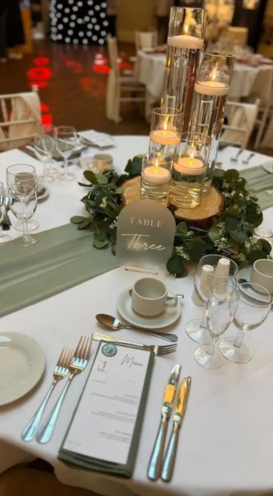 Round table with a sage green runner, floating candles in glass cylinders, eucalyptus and a Table Three sign