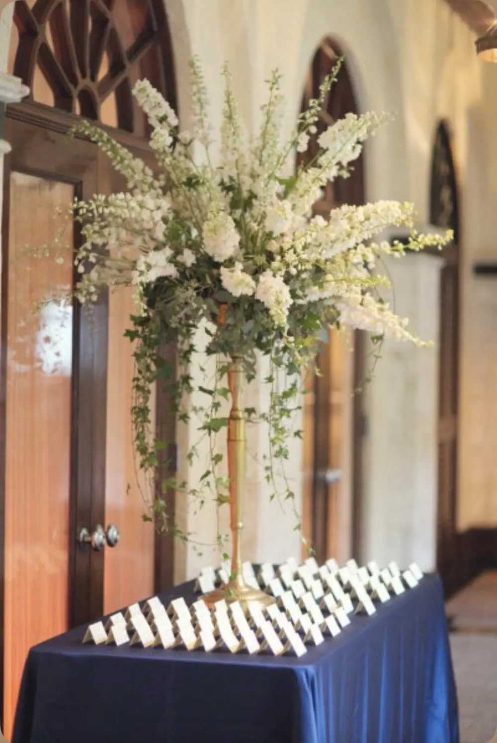 Tall white delphinium arrangement on a brass stand above an escort card table