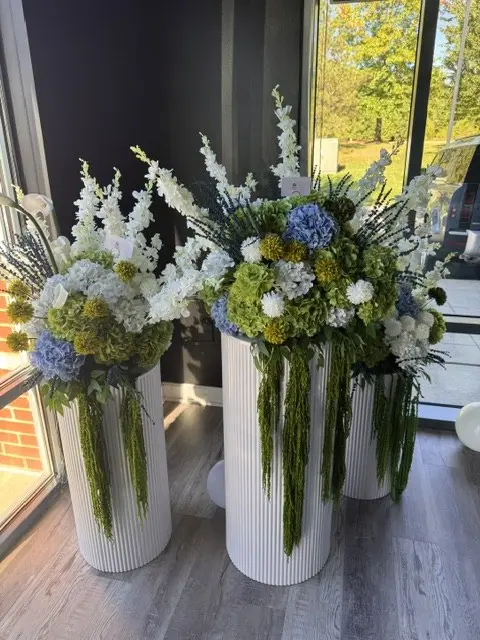 Three white fluted pedestals topped with blue and green hydrangea, white delphinium and trailing greenery