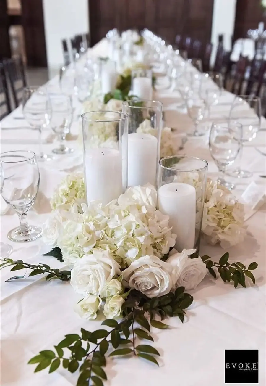White hydrangea and rose centerpiece ringed with greenery and pillar candles on a long table