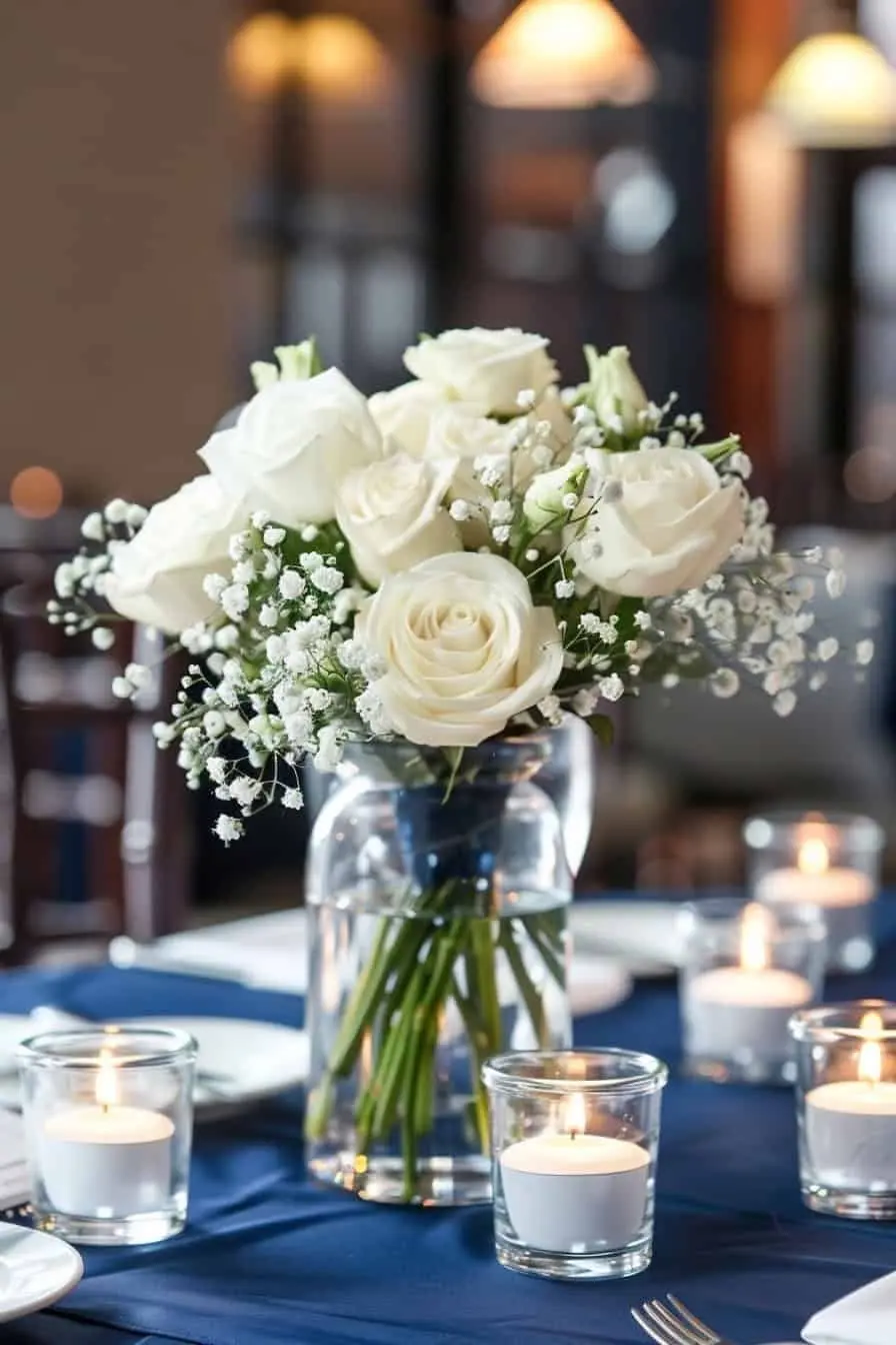 White roses and baby's breath in a glass vase on a navy tablecloth with votive candles